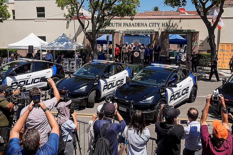Police cars parked in front of South Pasadena City Hall as journalists and a crowd film a podium at a daytime press conference.