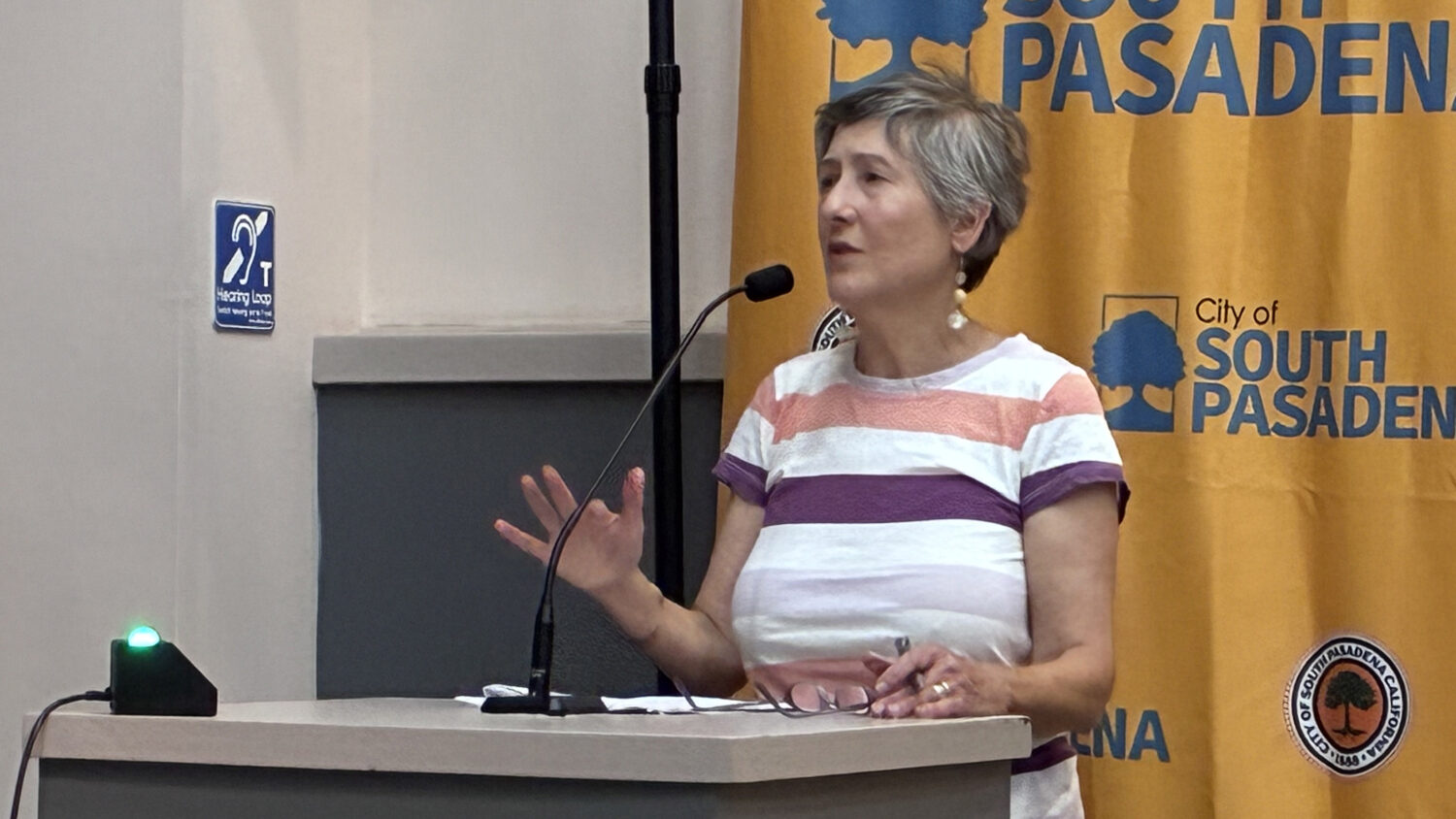 Bianca Richards standing at a podium giving public comment with a microphone in front of a yellow banner reading'City of South Pasadena' and a tree logo behind her.