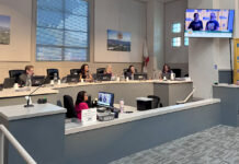 South Pasadena City council chamber: City Councilmember officials seated at a curved desk with laptops, microphones, and nameplates; a man stands at a podium on the right assessing a presentation on a wall screen.