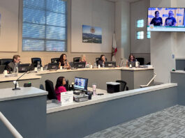South Pasadena City council chamber: City Councilmember officials seated at a curved desk with laptops, microphones, and nameplates; a man stands at a podium on the right assessing a presentation on a wall screen.