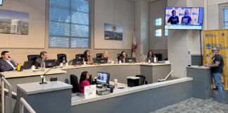 South Pasadena City council chamber: City Councilmember officials seated at a curved desk with laptops, microphones, and nameplates; a man stands at a podium on the right assessing a presentation on a wall screen.