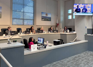South Pasadena City council chamber: City Councilmember officials seated at a curved desk with laptops, microphones, and nameplates; a man stands at a podium on the right assessing a presentation on a wall screen.