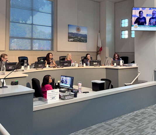 South Pasadena City council chamber: City Councilmember officials seated at a curved desk with laptops, microphones, and nameplates; a man stands at a podium on the right assessing a presentation on a wall screen.
