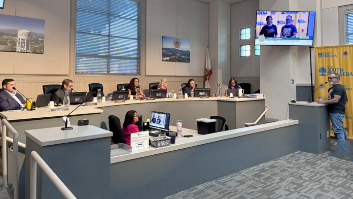 South Pasadena City council chamber: City Councilmember officials seated at a curved desk with laptops, microphones, and nameplates; a man stands at a podium on the right assessing a presentation on a wall screen.