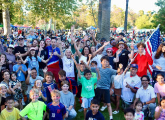 South Pasadena World Cup in Garfield Park crowd at a park celebration, many people raising hands and smiling, with an American flag visible in the background.