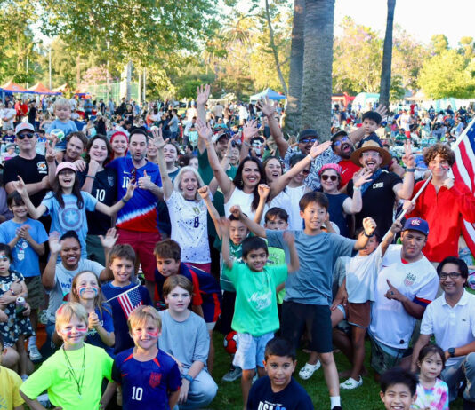 South Pasadena World Cup in Garfield Park crowd at a park celebration, many people raising hands and smiling, with an American flag visible in the background.