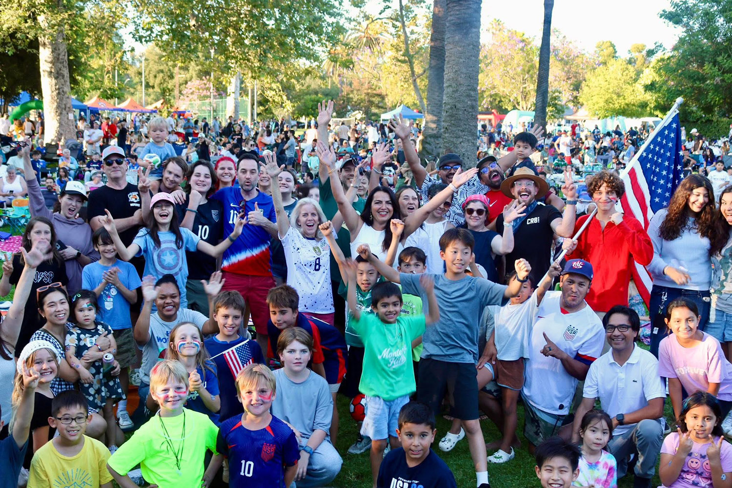 South Pasadena World Cup in Garfield Park crowd at a park celebration, many people raising hands and smiling, with an American flag visible in the background.