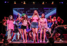 A group of teenagers dancing on a stage, centered around three girls in pink tops and skirts, with a large screen backdrop behind them.