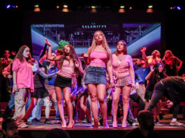 A group of teenagers dancing on a stage, centered around three girls in pink tops and skirts, with a large screen backdrop behind them.
