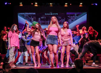 A group of teenagers dancing on a stage, centered around three girls in pink tops and skirts, with a large screen backdrop behind them.