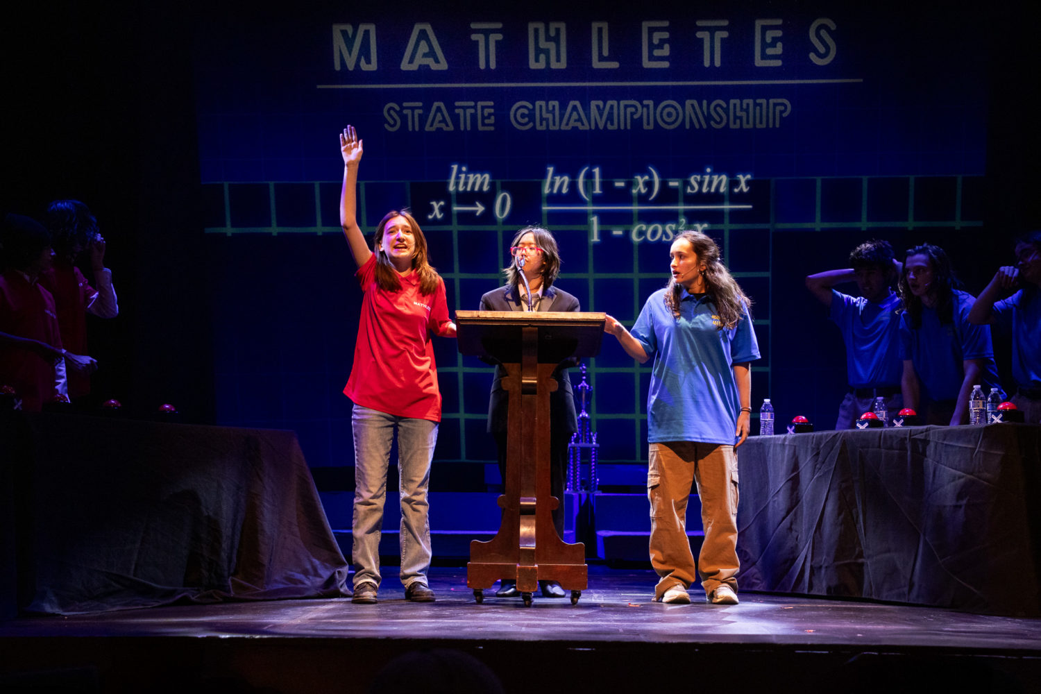 | The South Pasadenan | South Pasadena News Three students on stage at the Mathletes State Championship: left student in red raises hand, center speaks at the podium, right student in blue watches.