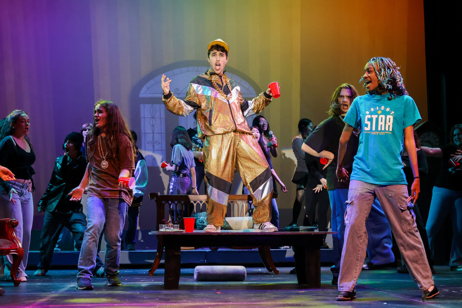 | The South Pasadenan | South Pasadena News Person in shiny gold tracksuit stands on a table center stage, arms raised, performing with a red cup in hand amid a colorful dance troupe.