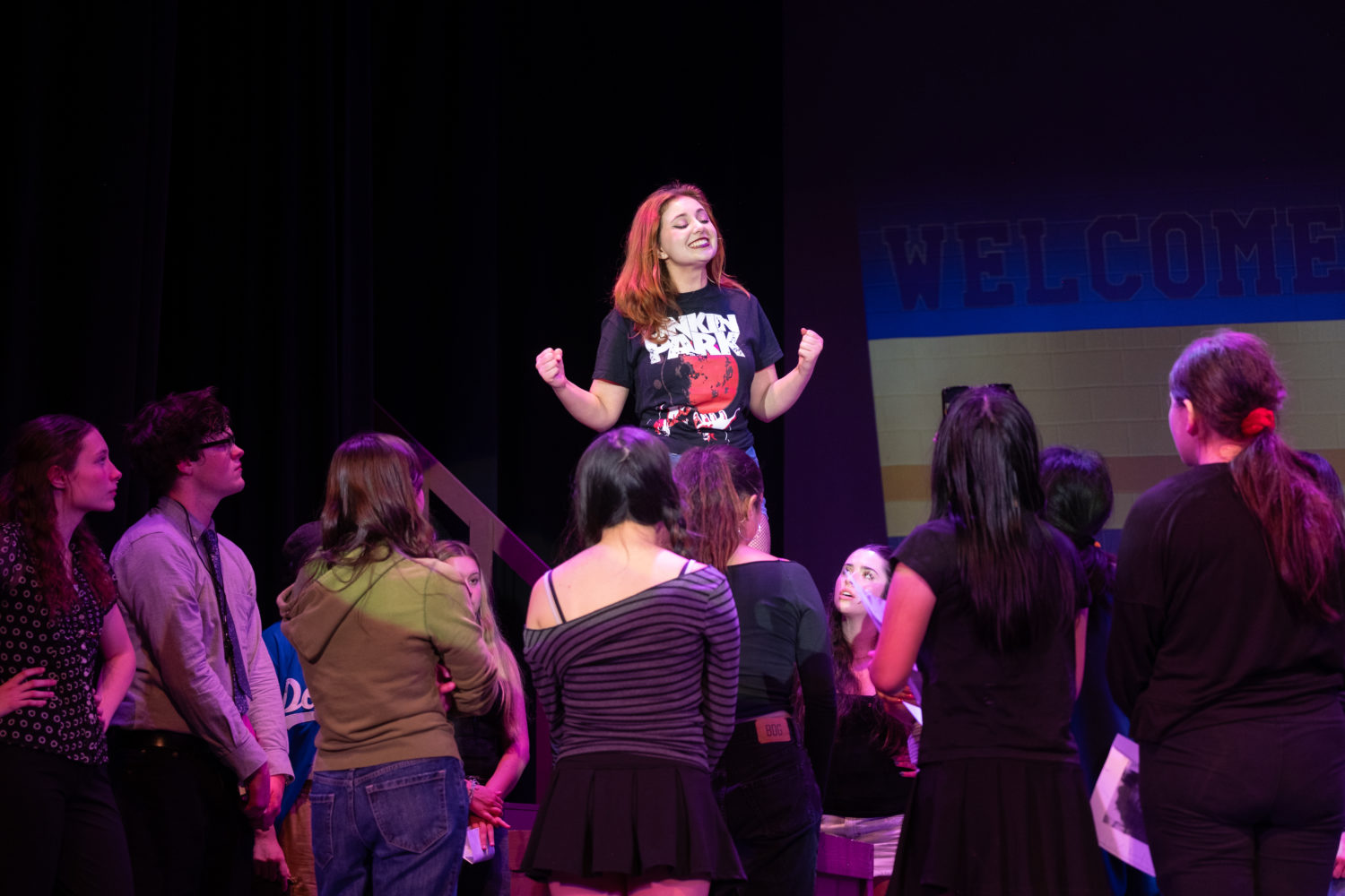 Girl on a stage with fists raised, addressing a circle of students in a dimly lit auditorium with a Welcome banner in the background.] ,