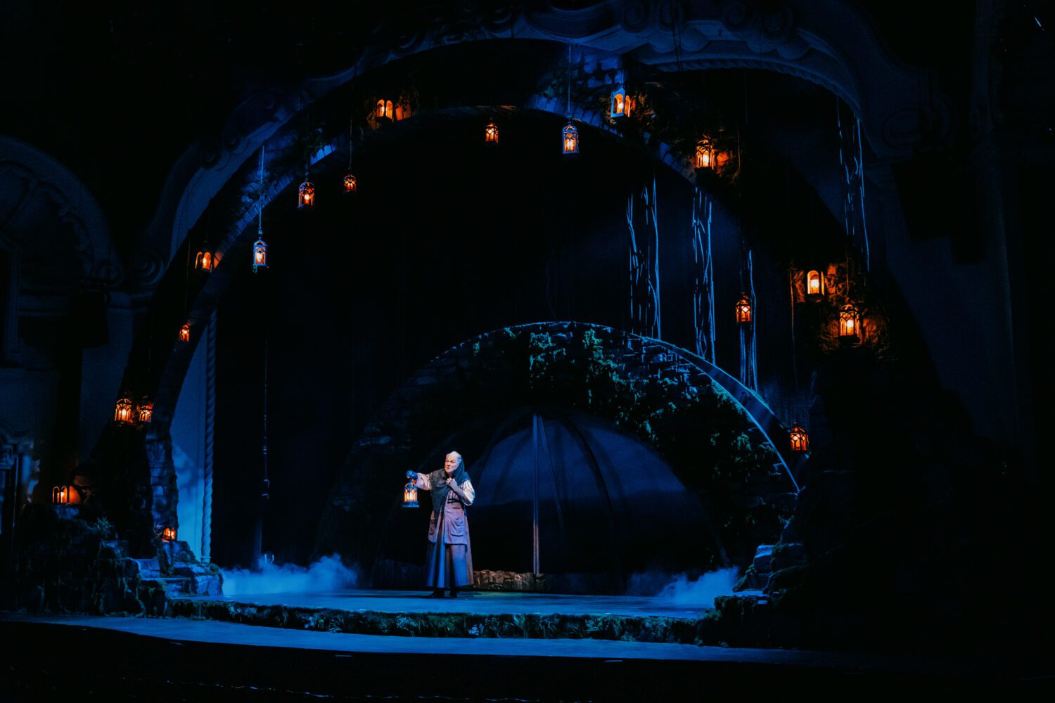 Performer (Tyne Daly) in a long robe stands center stage, holding a lantern under blue, moody lighting with circular arches and hanging lanterns San a misty atmosphere