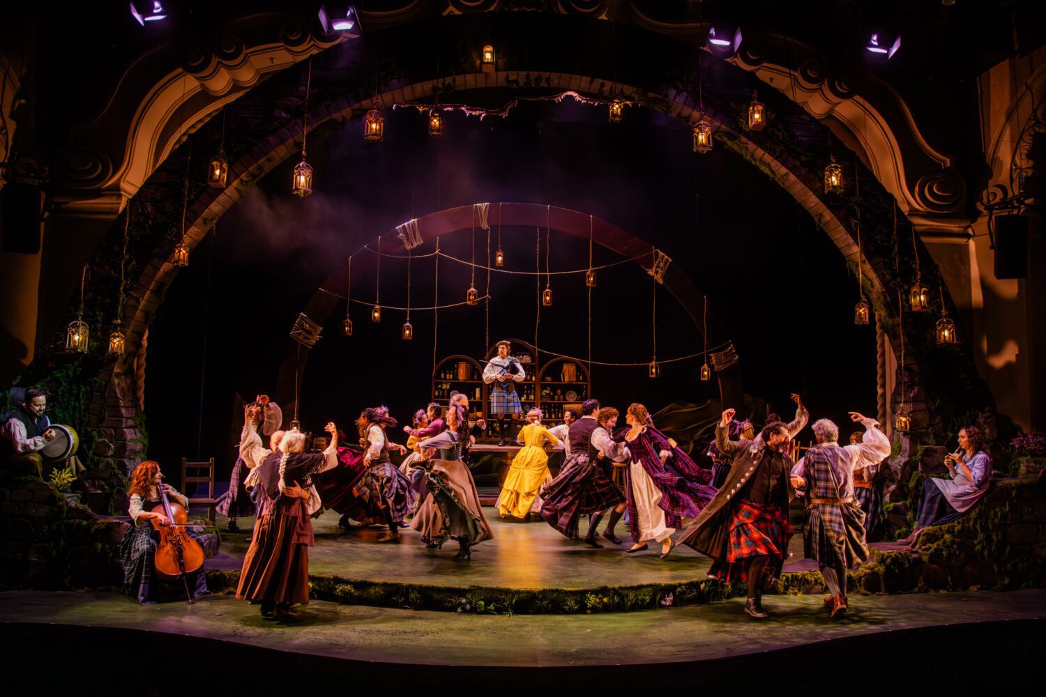 A lively stage scene with performers in 19th-century costumes dancing in a circle under hanging lanterns on a theater set.