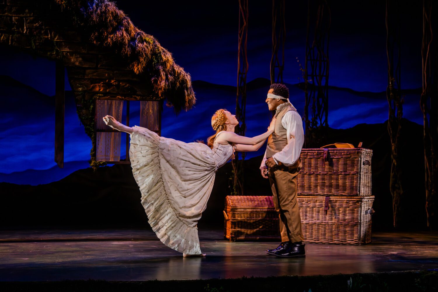 Woman in a flowing white gown kicks her leg up toward a man in a vest on a theater stage, with wicker baskets in the background.
