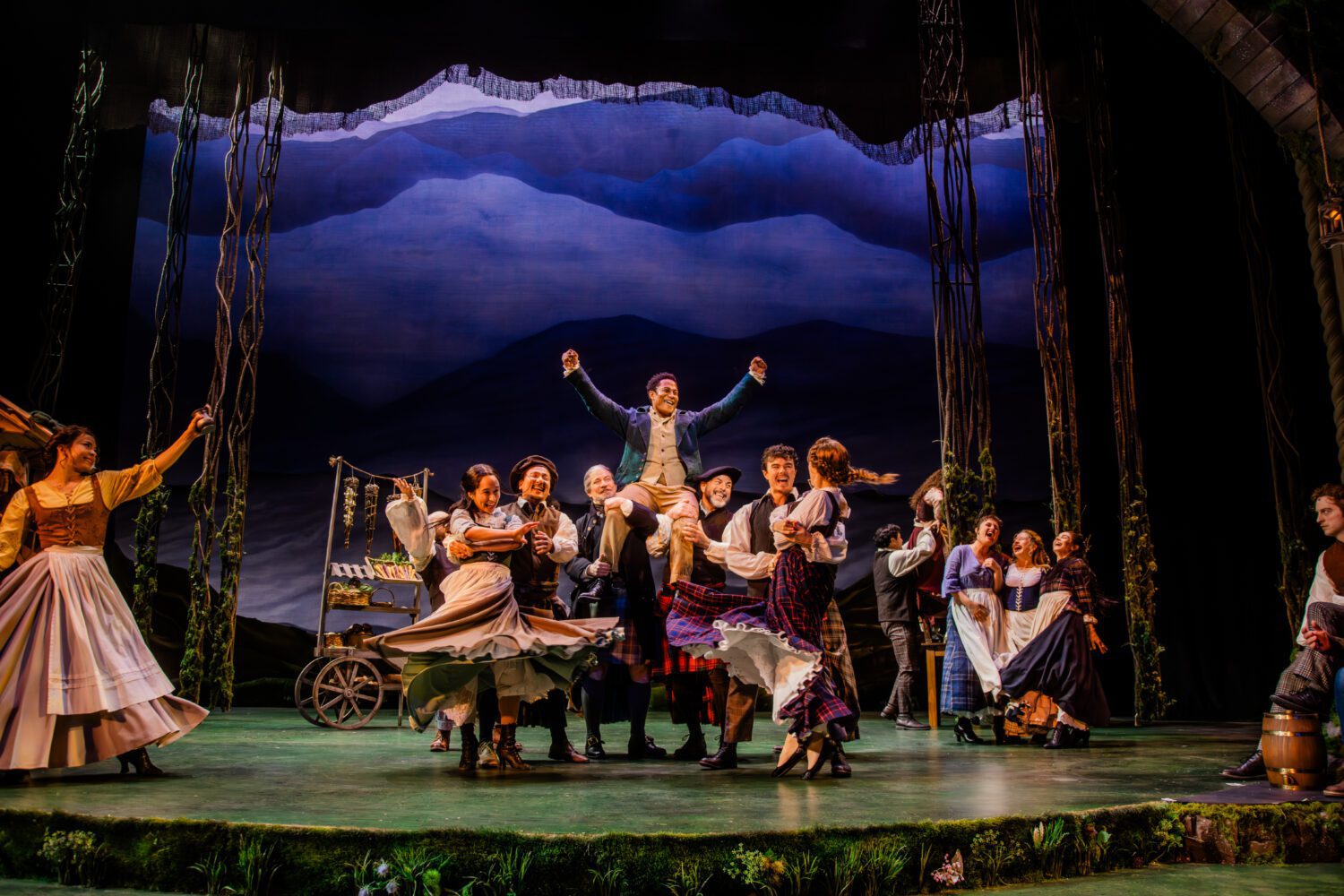 A lively theater scene with performers in period costumes cheering as a man in a blue coat is lifted atop the crowd center stage.