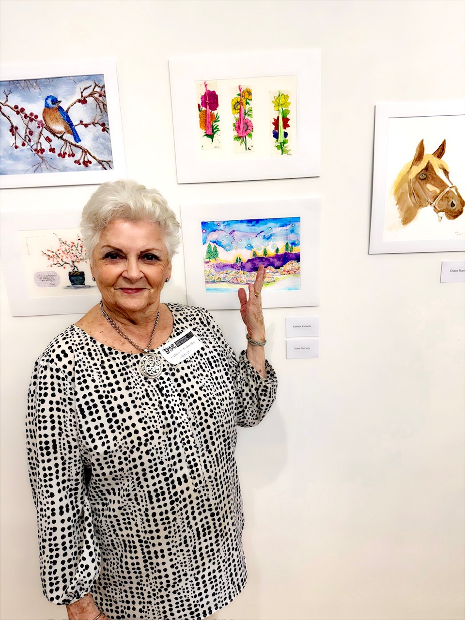 | The South Pasadenan | South Pasadena News Older woman with short white hair in a black-and-white polka-dot blouse stands beside framed artworks in a gallery, smiling at the camera.