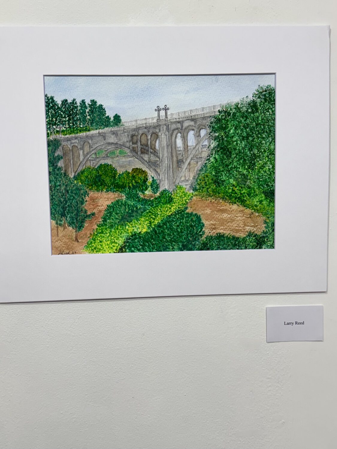 south-pasadena-news-07-12-2026-the-creative-life-blooms-senior-watercolorists 5 | The South Pasadenan | South Pasadena News Painting of a stone arch bridge with multiple arches, set in a lush green landscape under a blue sky, framed and signed by Larry Reed.