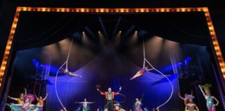 Circus stage with colorful performers; two aerialists swing from hoops above a central troupe under bright spotlights.