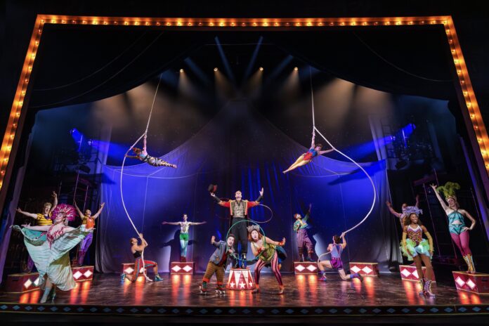 Circus stage with colorful performers; two aerialists swing from hoops above a central troupe under bright spotlights.