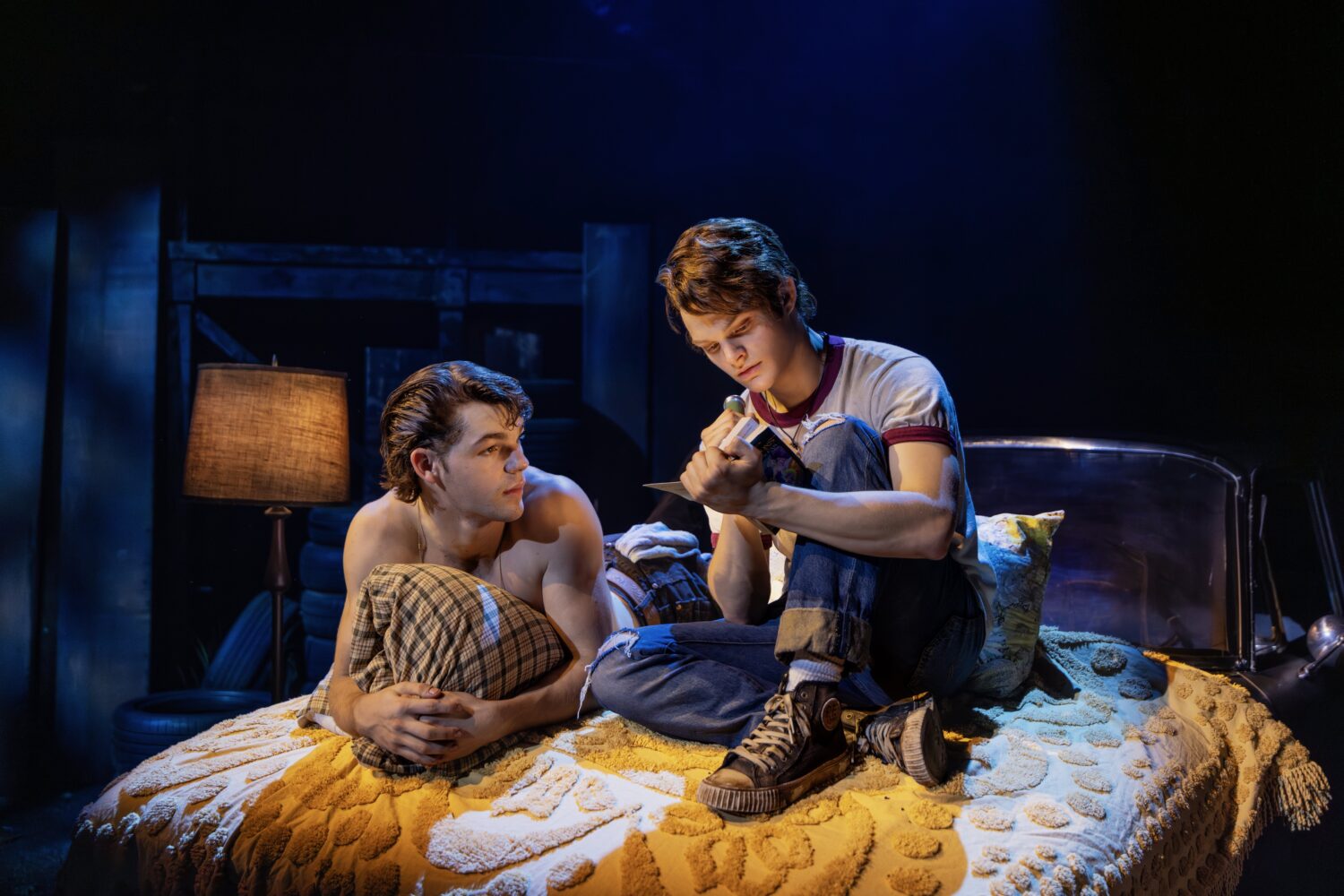 Two young men sit on a bed in a dimly lit room, one shirtless with plaid pants and the other in jeans, reading a notebook together.