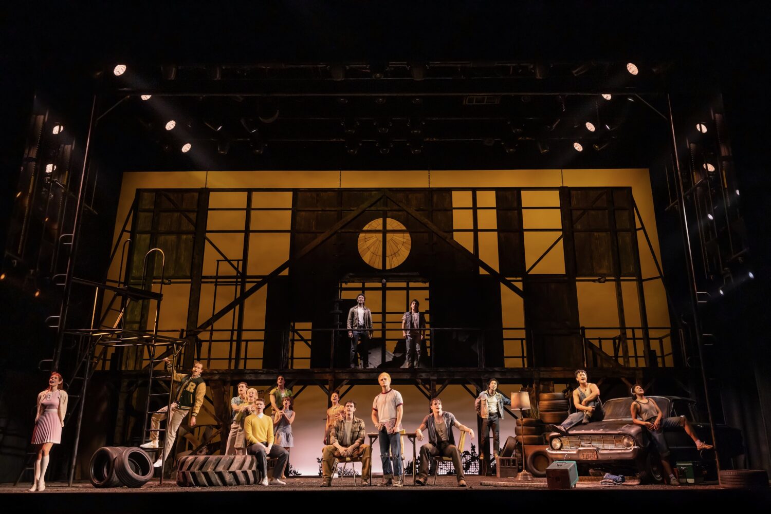 A group of actors on a two-level stage set with a yellow-lit industrial backdrop, a car and tires on the right, and performers in varied poses in the foreground.