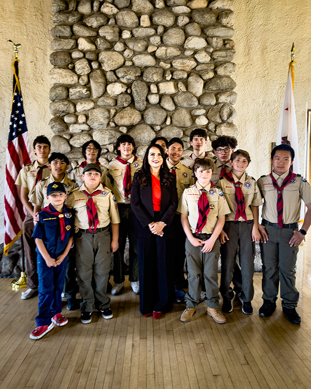Group of scouts in tan uniforms with a female leader in a red blouse, posing indoors in front of a stone fireplace and Flags visible on either side.